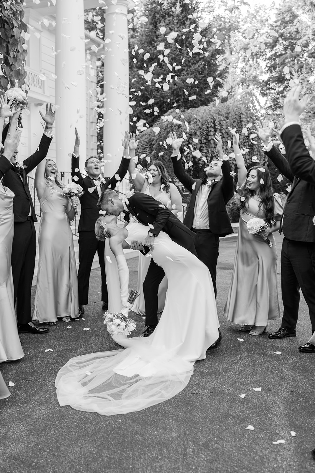 Black and white photo of the groom dipping and kissing the bride as the wedding party throws rose petals.